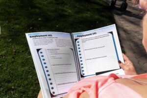 woman in pink reading "employment 101" workbook pages with grassy lawn in background